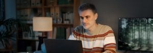 Un bărbat purtând un pulover în dungi stă la birou și se uită la laptopul din fața lui