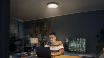 Un bărbat stă la un birou cu un laptop, iluminat de sus de o lampă rotundă de plafon